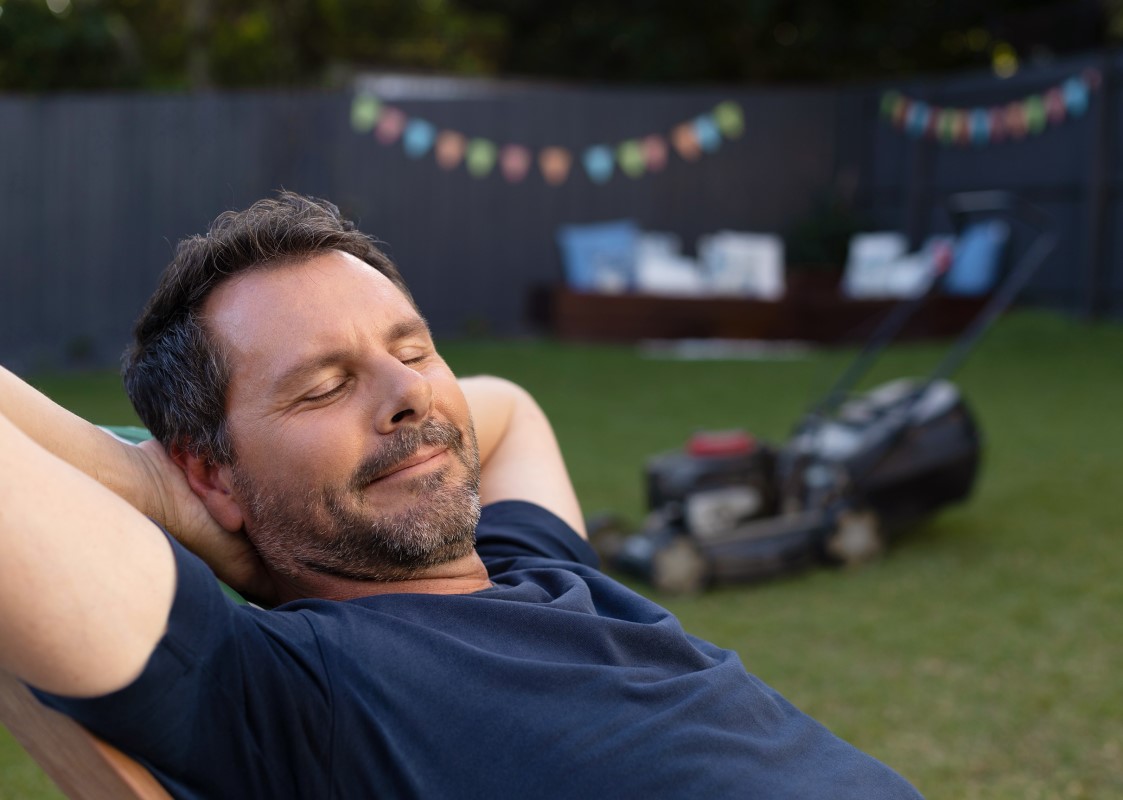 Man lying in deck chair with eyes closed with a lawn mower in the background