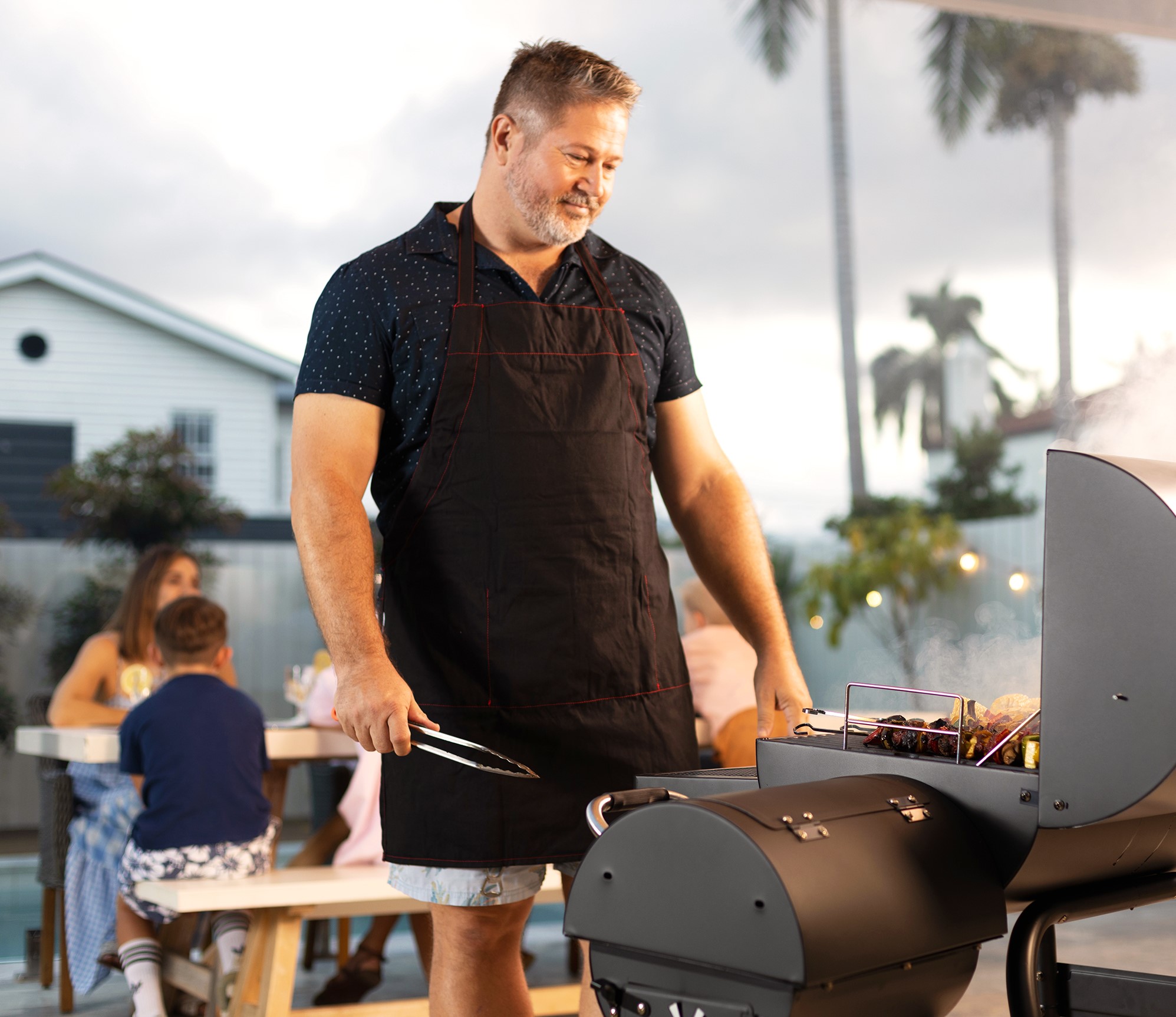 Man in black shirt grilling on a BBQ with family members seated in the background