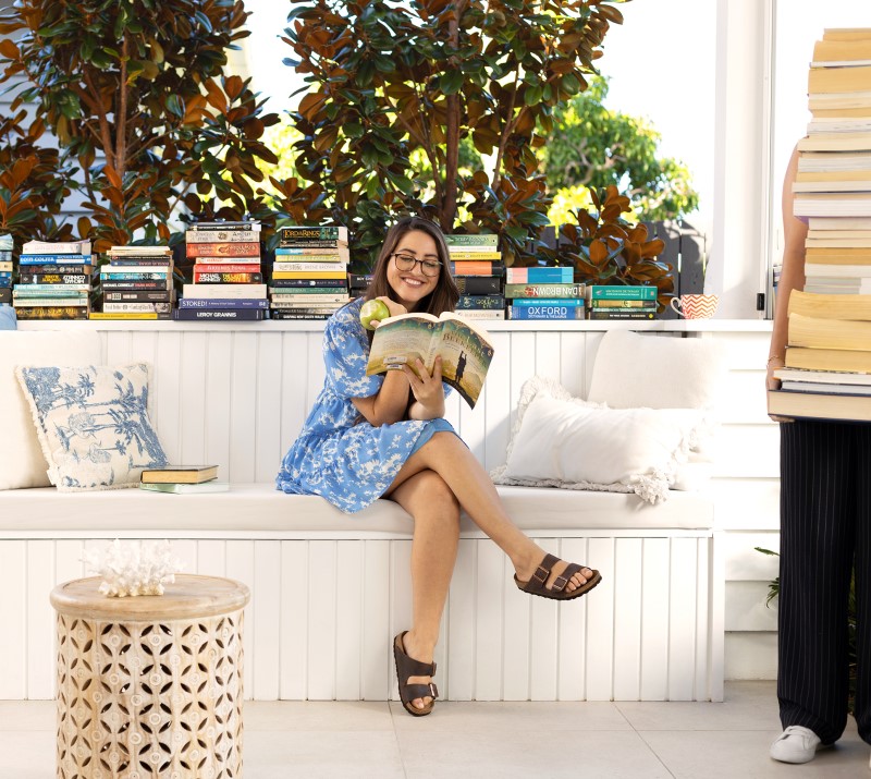 Woman in a blue dress seated outdoors reading a book and eating a patio while someone holds a large stack of books nearby