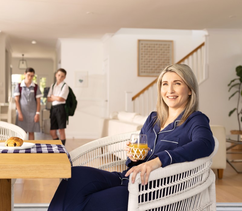 A woman in navy pyjamas relaxes with a glass of juice while two children in school uniforms look on in the background